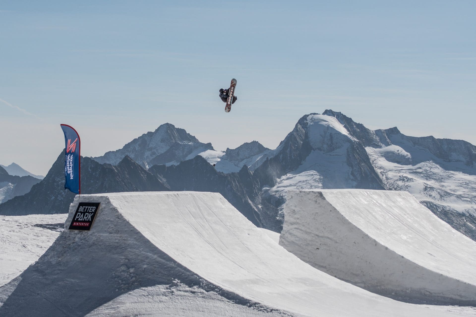 Snowboarder flying over a park jump with alpine peaks behind
