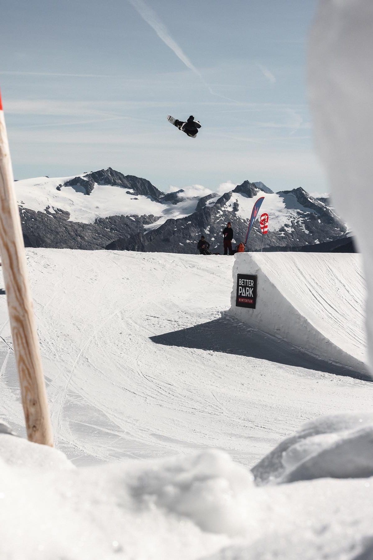 Snowboarder spinning above a park jump with snowy peaks behind