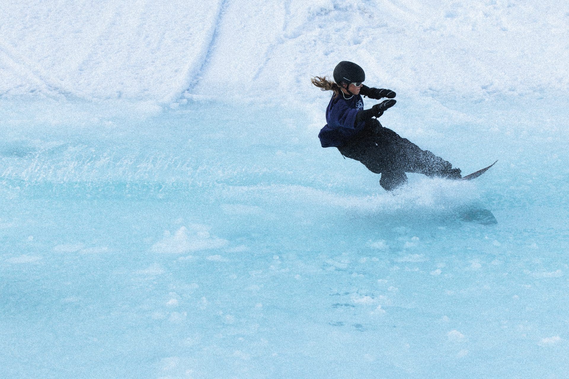 Snowboarder Selin Lakatha skimming across a turquoise pond slide, spray flying behind her board